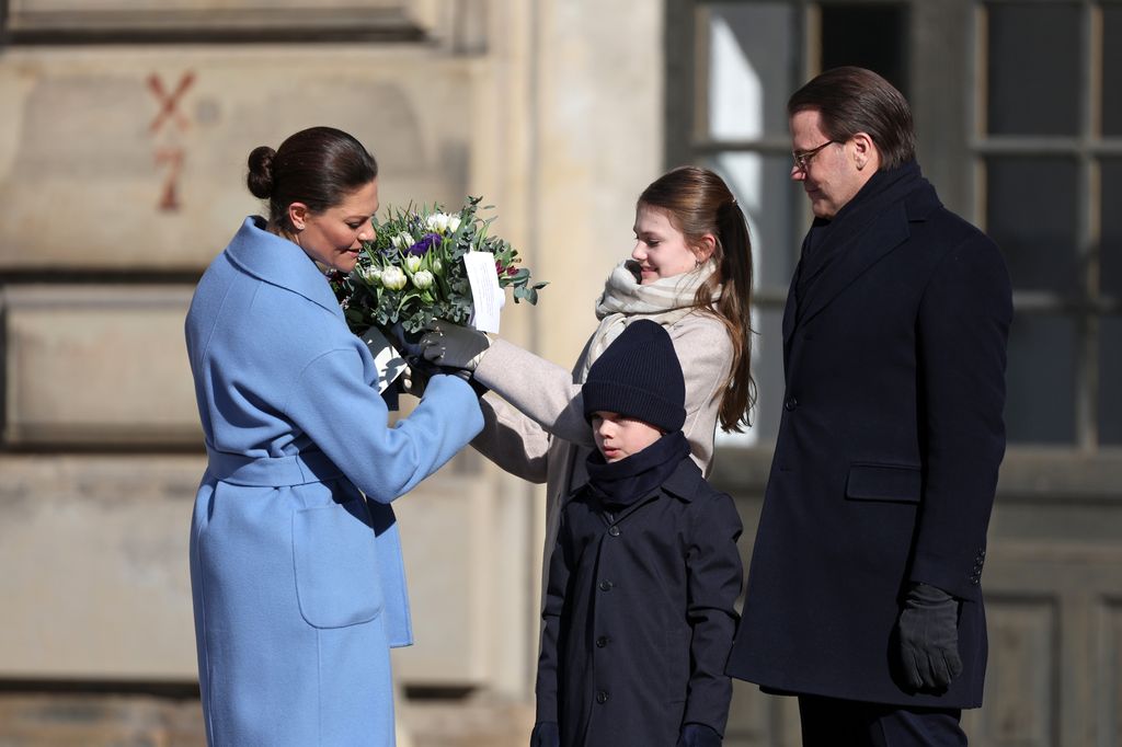 Princesa herdeira Victoria com casaco azul celeste entregando flores para Estelle