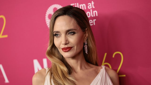 Angelina Jolie with long, wavy blonde hair is smiling slightly. She is in a white, elegant pleated gown. Her makeup includes bold red lipstick, and she is standing against a vibrant pink backdrop.