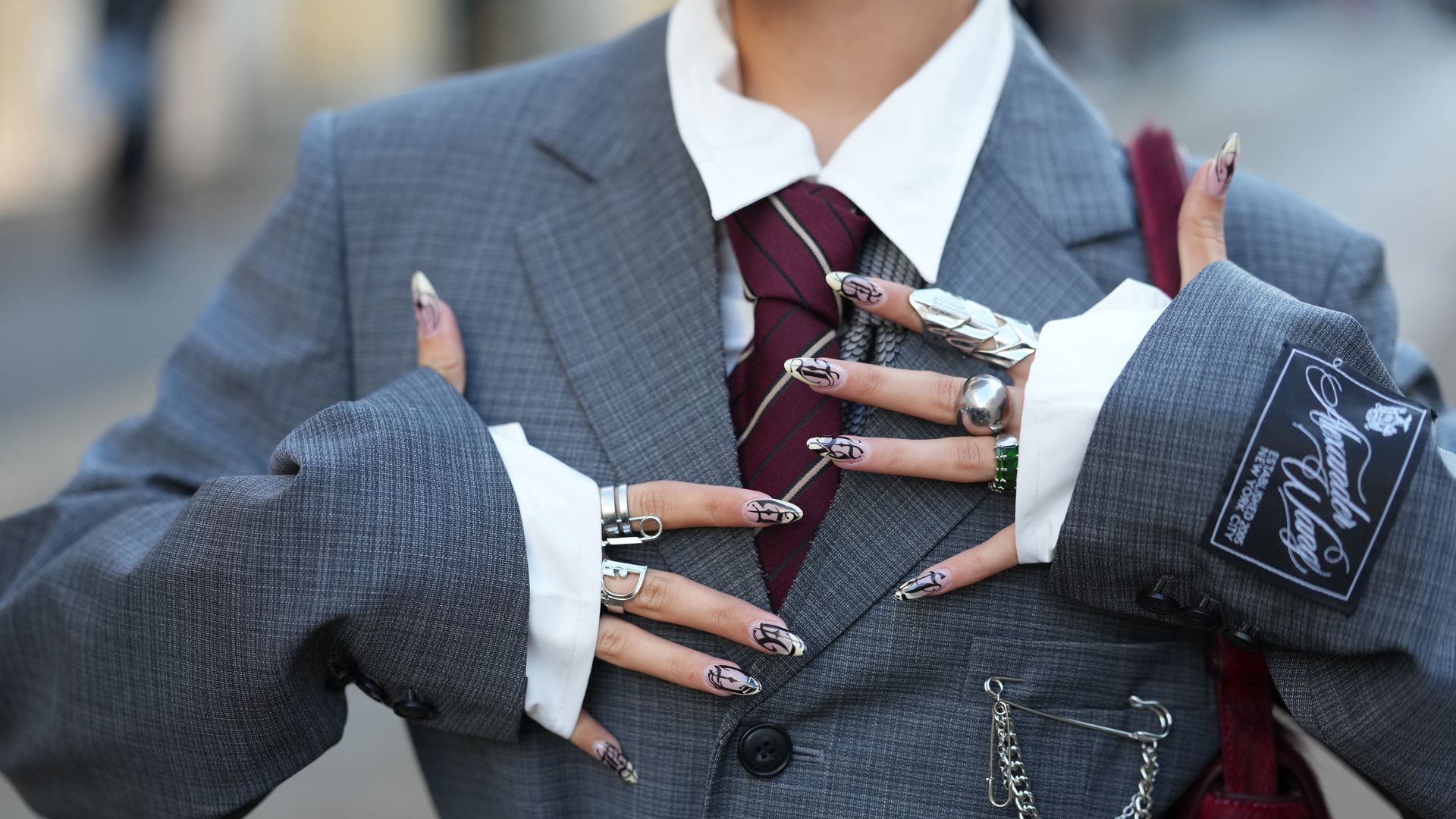 MILAN, ITALY - SEPTEMBER 25: A guest wears large multiple silver rings, a burgundy fuzzy shoulder bag, a white button-down shirt with visible collar, a burgundy and navy striped tie, a charcoal grey checked wool suit jacket with notch lapels, front flap pockets, a black sleeve patch and a silver chain draped from the pocket and worn as a belt, outside MM6, during Milan Fashion Week - Womenswear Spring/Summer 2026, on September 25, 2025 in Milan, Italy (Photo by Edward Berthelot/Getty Images)
