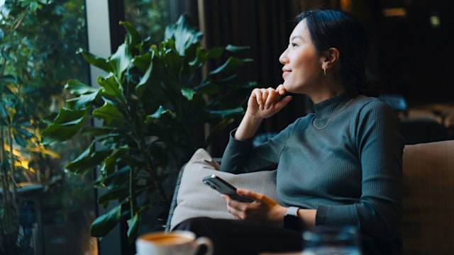 Confident young woman with smartphone looking out through window while sitting in a cafe having coffee.