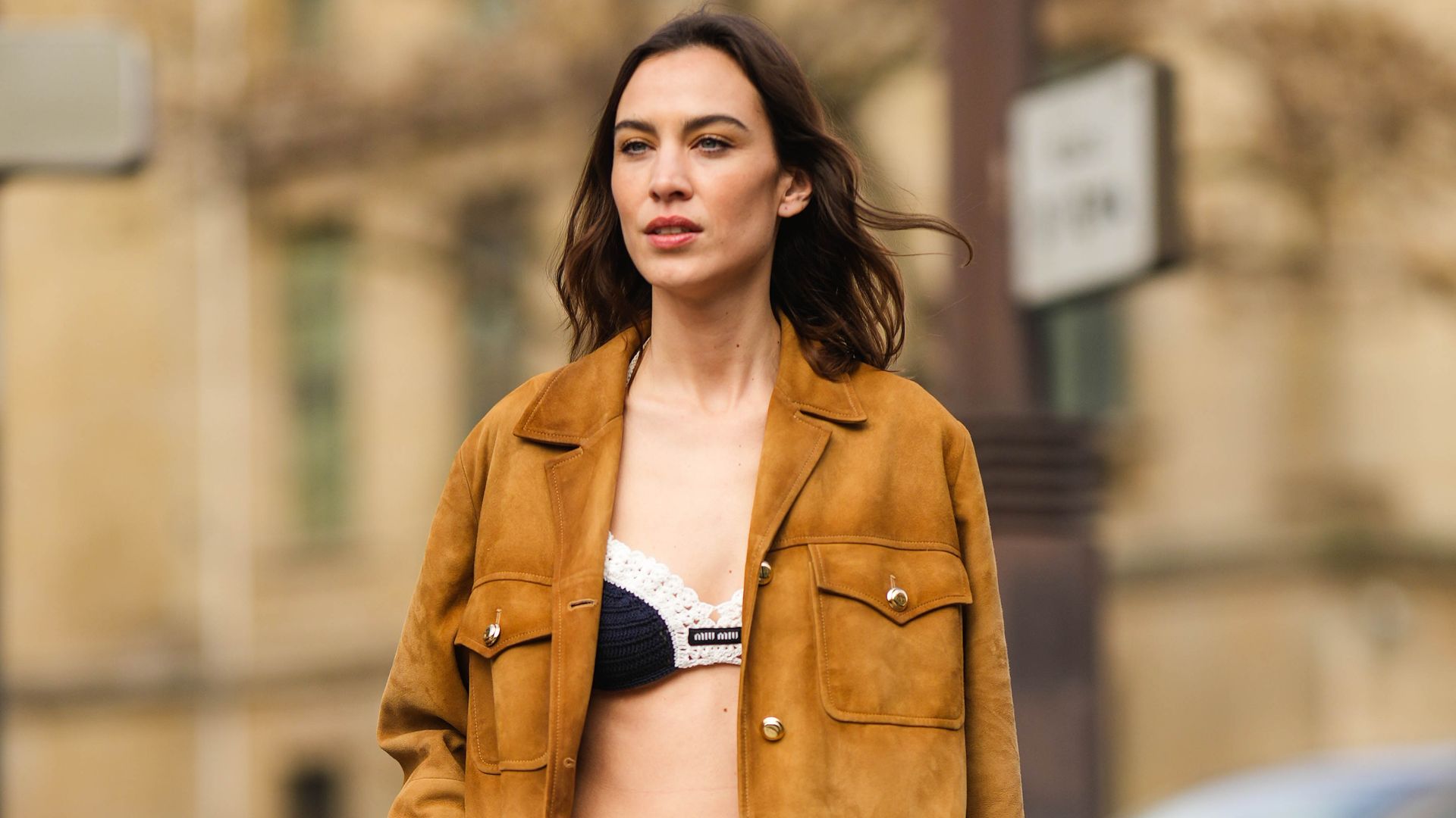 Alexa Chung wears a black braided wool with embroidered white borders V-neck cropped bra underwear from Miu Miu, a camel suede buttoned jacket, a white pleated / accordion short skirt from Miu Miu, a black shiny leather belt, a pale gray embossed pattern handbag from Miu Miu, black shiny leather pointed loafers from Miu Miu