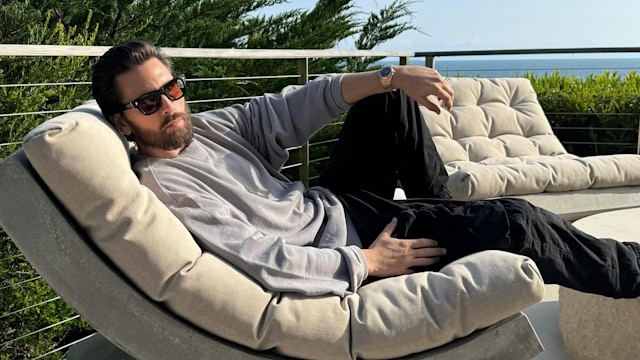 Scott Disick lying on an outdoor seat in the sun wearing a grey top