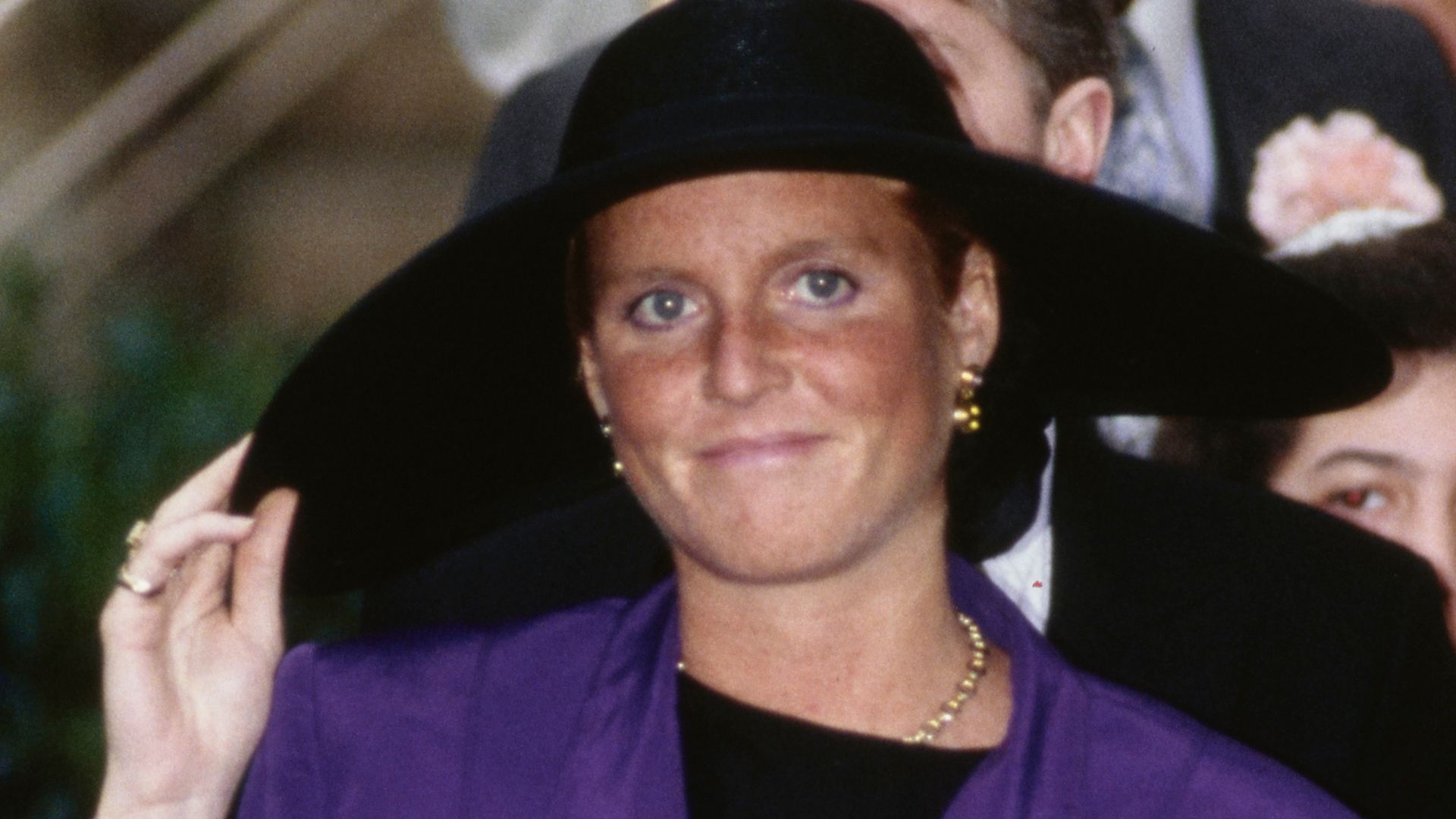 Sarah, Duchess of York wearing a baby bump-skimming purple outfit and holding her hat at the wedding of the Marquis of Marlborough and Rebecca Few Brown