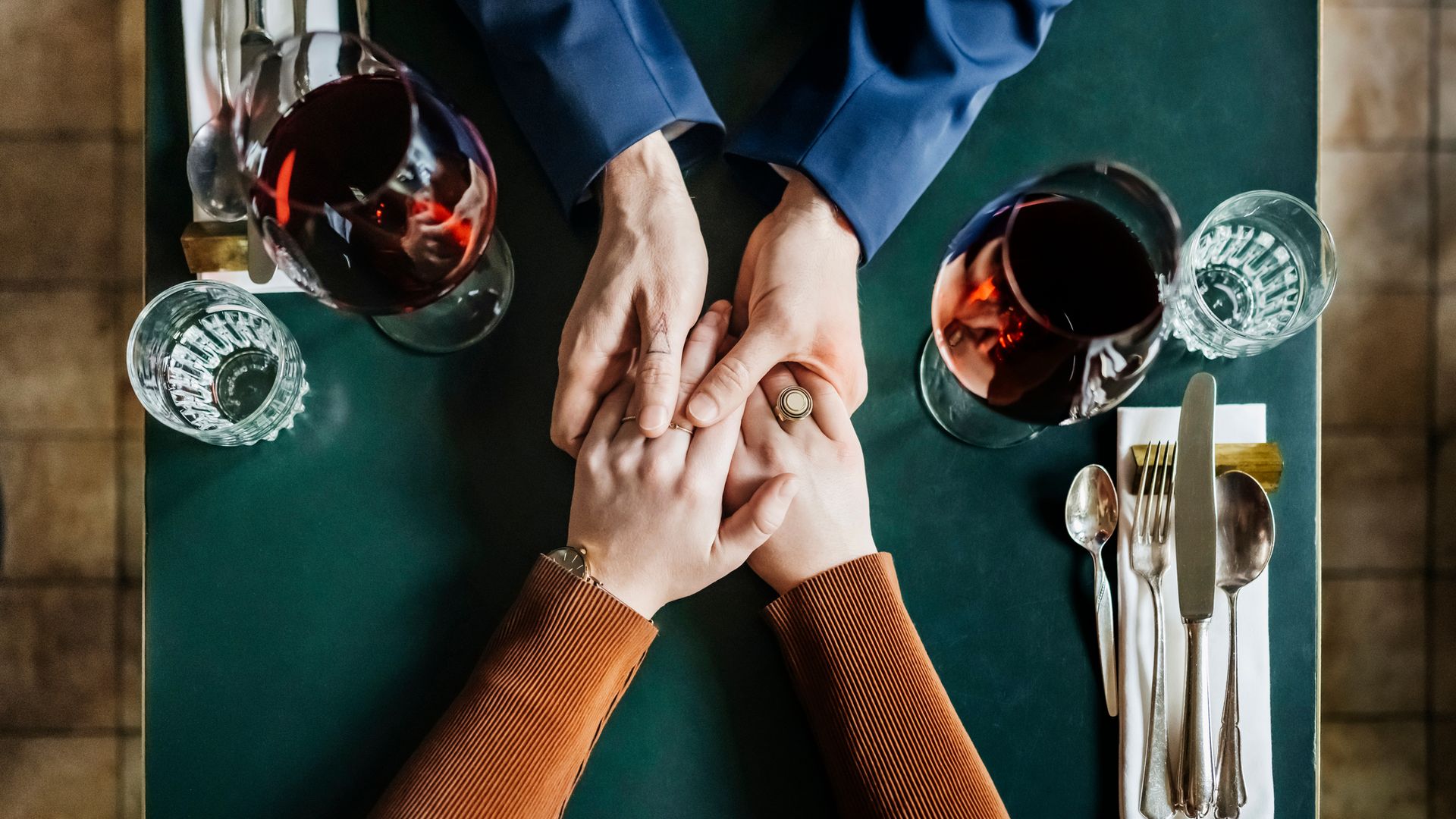 An aerial view of a couple holding hands and drinking red wine while sitting at a restaurant table for lunch together.