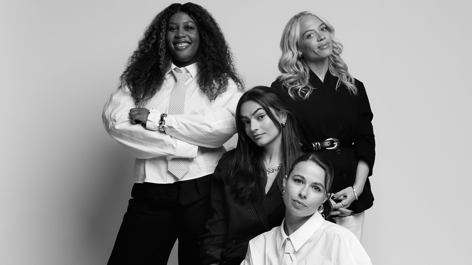 Black-and-white studio portrait of four women posing together in tailored outfits, sitting and standing in a minimal backdrop.