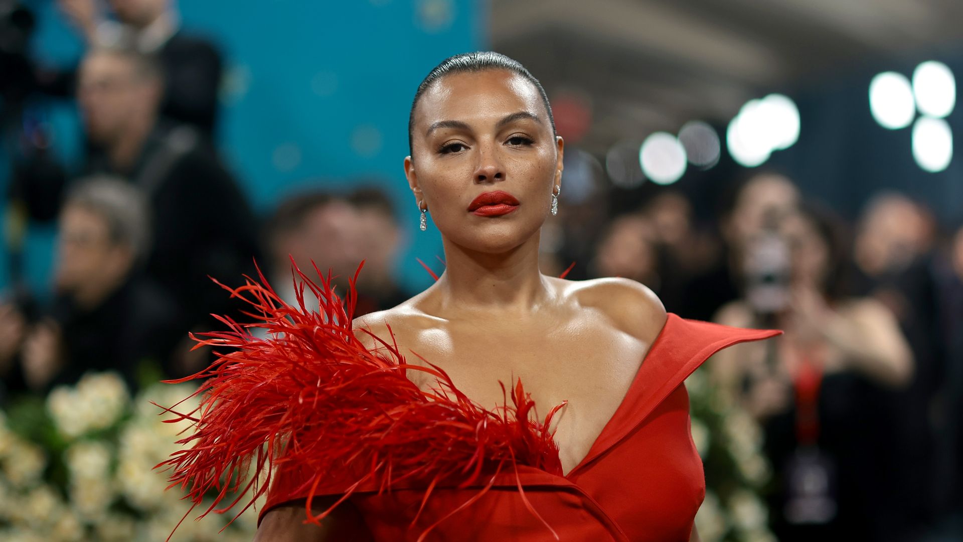 Paloma Elsesser at the 2025 Met Gala wearing a red gown with a feathered shoulder and off-the-shoulder neckline, striking a confident pose.