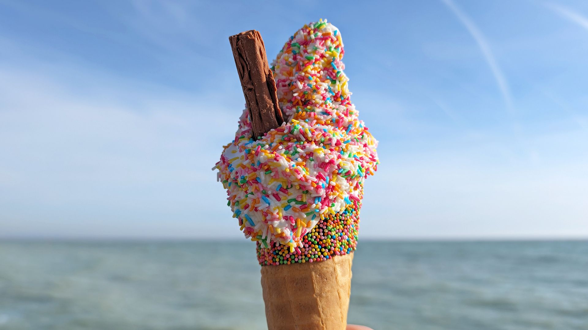 hand holding ice cream cone with chocolate flake and rainbow sprinkles at seaside beach with blue sky and sea