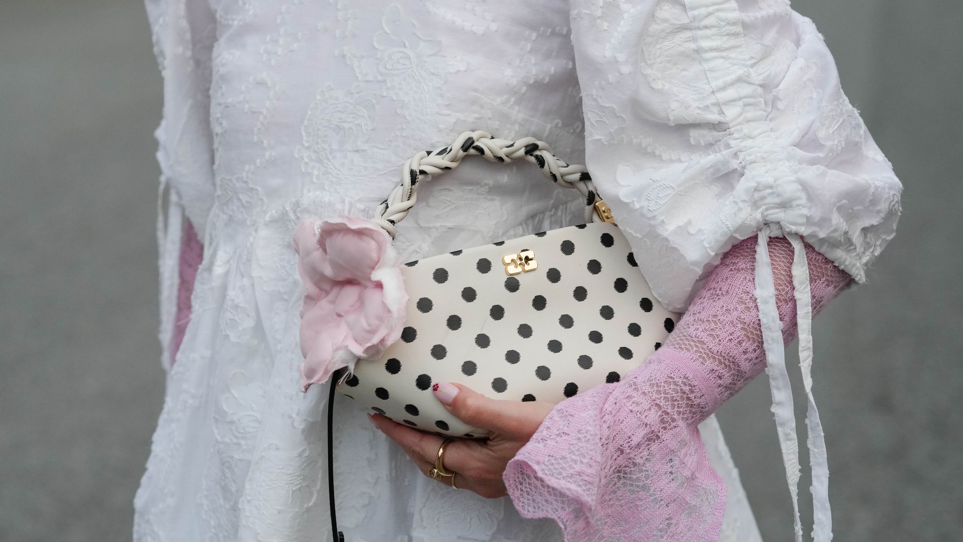  A guest wears a white dress in textured embroidered floral fabric with voluminous sleeves gathered by ruched drawstring seams and long ribbon ties, layered over sheer pink lace gloves with fluted, scalloped cuffs; a small cream leather top-handle bag with black polka dots, a braided cream-and-black handle, gold logo hardware, a pale pink fabric flower trim at the handle base, and a thin black shoulder strap; stacked gold rings including slim bands and a signet-style ring; and short nails painted with a sheer light polish and a tiny red accent at the tips, outside Rotate, during Copenhagen Fashion Week, on August 07, 2025 in Copenhagen, Denmark