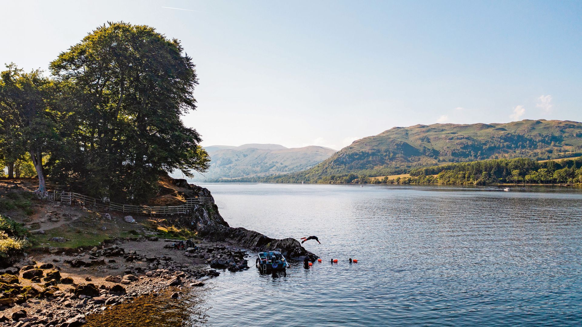 A tranquil lakeside scene with a rocky shore, trees, and distant hills under a clear blue sky. Swimmers and a small boat are visible near the water’s edge.