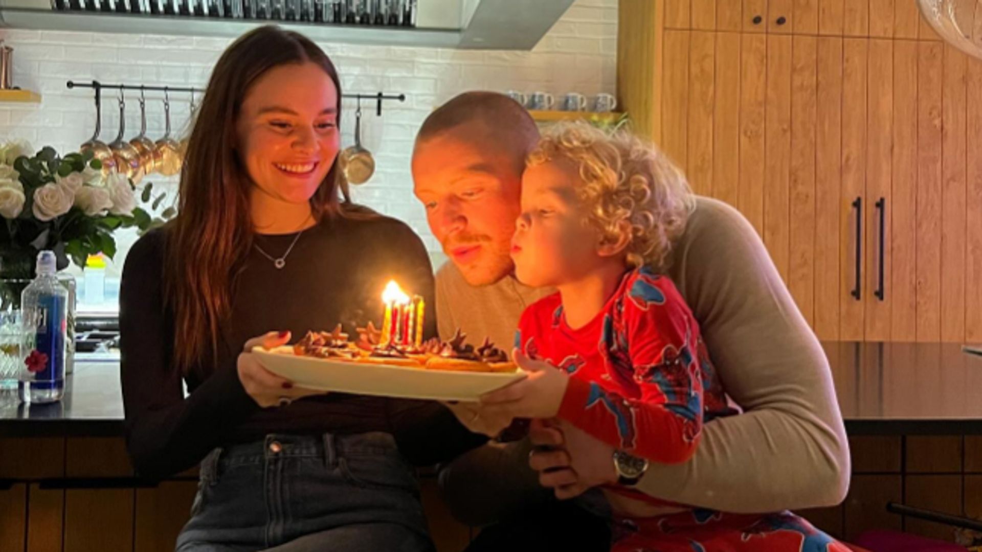 Holly Ramsay holding a birthday cake while Adam Peaty and his son blow out the candles