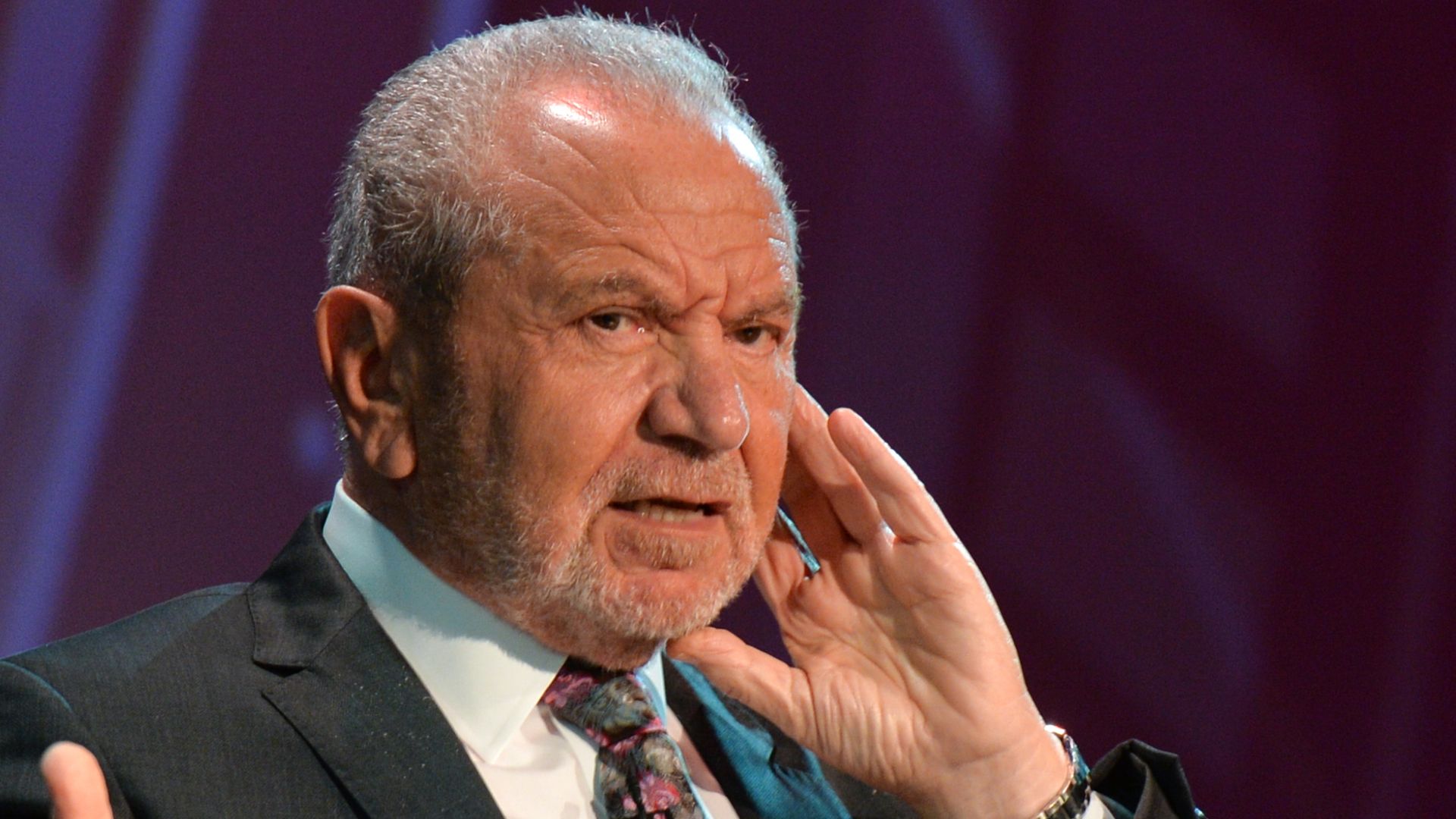 alan sugar speaking on stage in suit and tie 