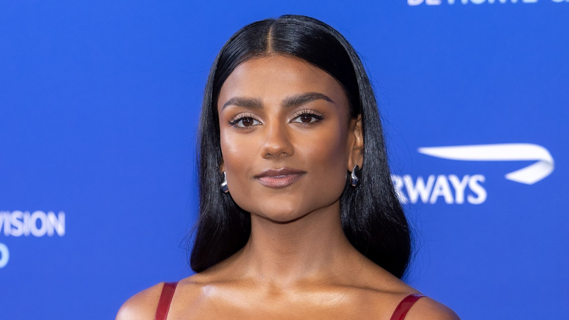 Simone Ashley stands on a navy blue carpet against a backdrop reading "63rd Festival de Télévision de Monte-Carlo." She is wearing a fitted, glossy red dress with thin shoulder straps and a unique balloon-style skirt that cinches in at the hem. The fabric has a highly reflective, lacquered texture that gives it a modern, high-shine appearance. Her long black hair is sleek and straight, parted in the middle. She pairs the dress with open-toe heels with transparent straps and clear block heels, giving her an almost floating appearance.