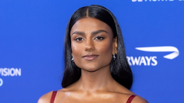 Simone Ashley stands on a navy blue carpet against a backdrop reading "63rd Festival de Télévision de Monte-Carlo." She is wearing a fitted, glossy red dress with thin shoulder straps and a unique balloon-style skirt that cinches in at the hem. The fabric has a highly reflective, lacquered texture that gives it a modern, high-shine appearance. Her long black hair is sleek and straight, parted in the middle. She pairs the dress with open-toe heels with transparent straps and clear block heels, giving her an almost floating appearance.