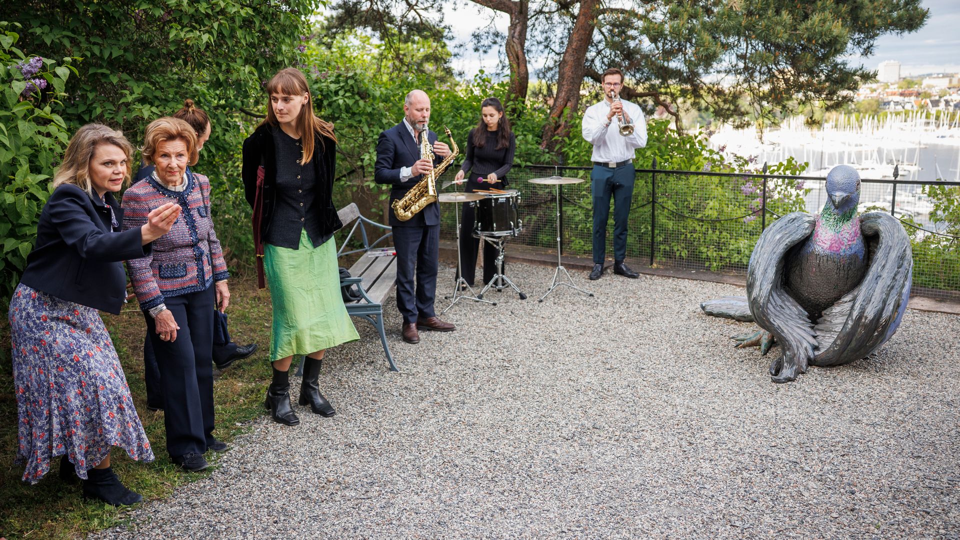 Queen Sonja and a group of people looking at a sculpture of a pidgeon while a small band plays