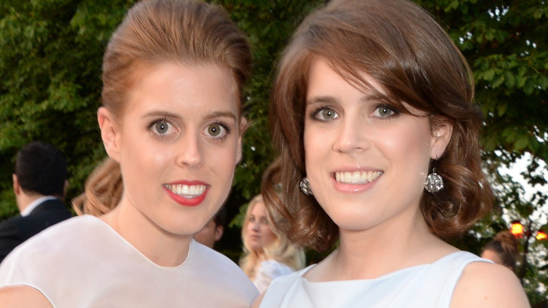 Princess Beatrice in a white dress and Princess Eugenie in a blue dress smiling outside at The Serpentine Gallery Summer Party in 2014