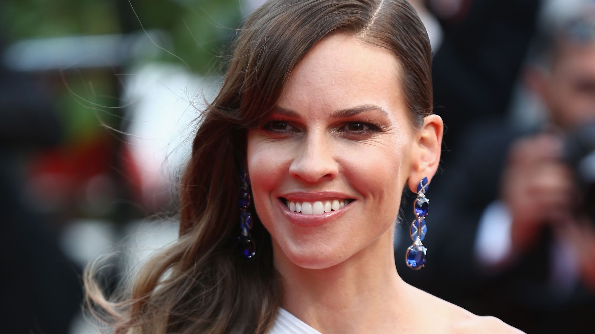 Hilary Swank in a one-shouldered gown and blue earrings smiling in an outdoor setting