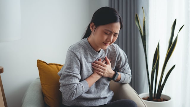 Asian woman suffering from chest pain breathing problems holding hands on her chest sitting on sofa in living room at home.