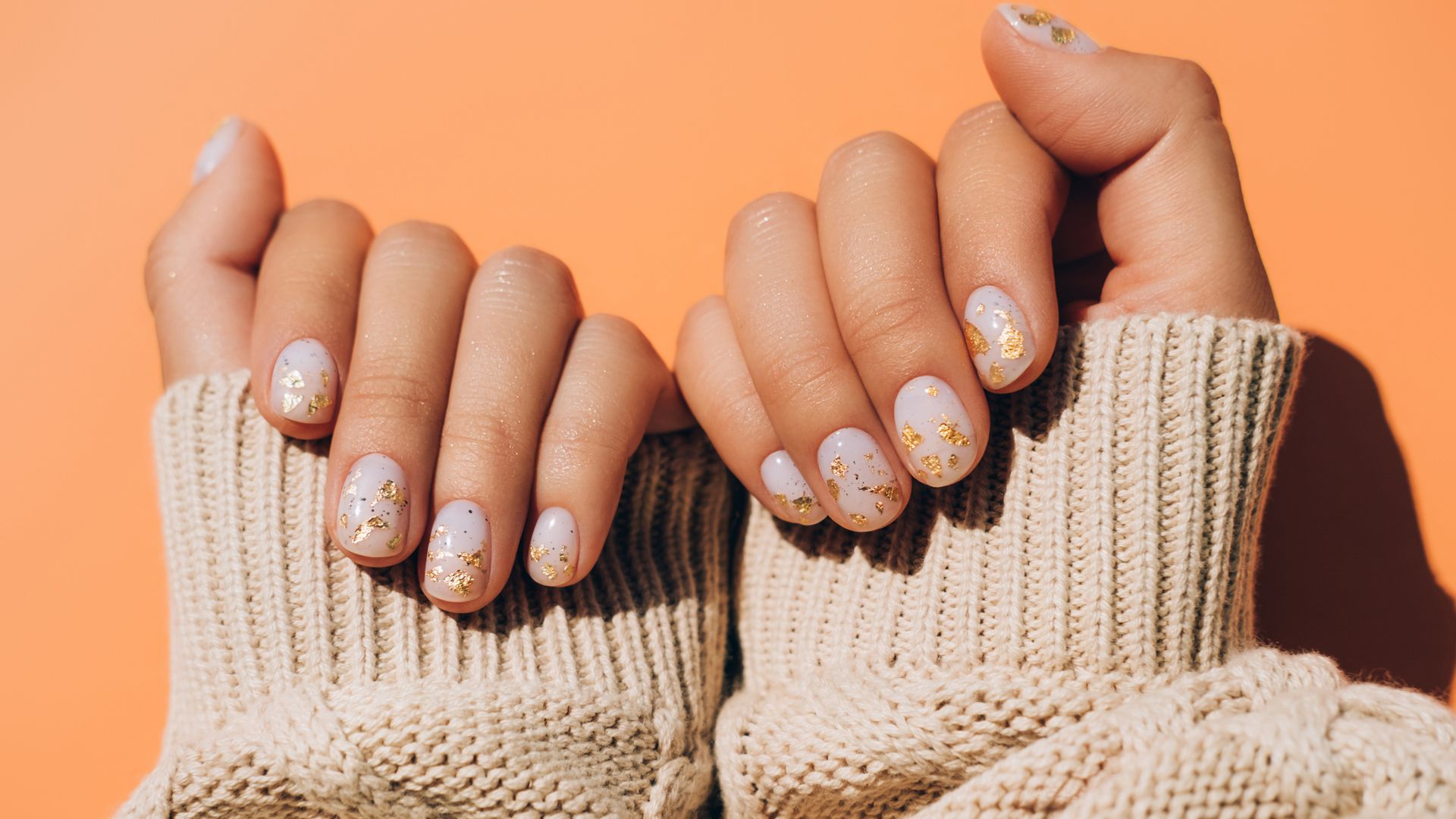 Glowing hands in sweater with knitted sleeves and with nude color manicure with gold particles on orange table. 