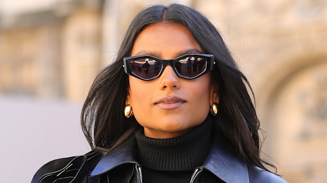 Simone Ashley wears a cropped leather jacket, black top and black mini skirt. She poses with one hand in her pocket and wears sunglasses and a small pair of gold hoop earrings.