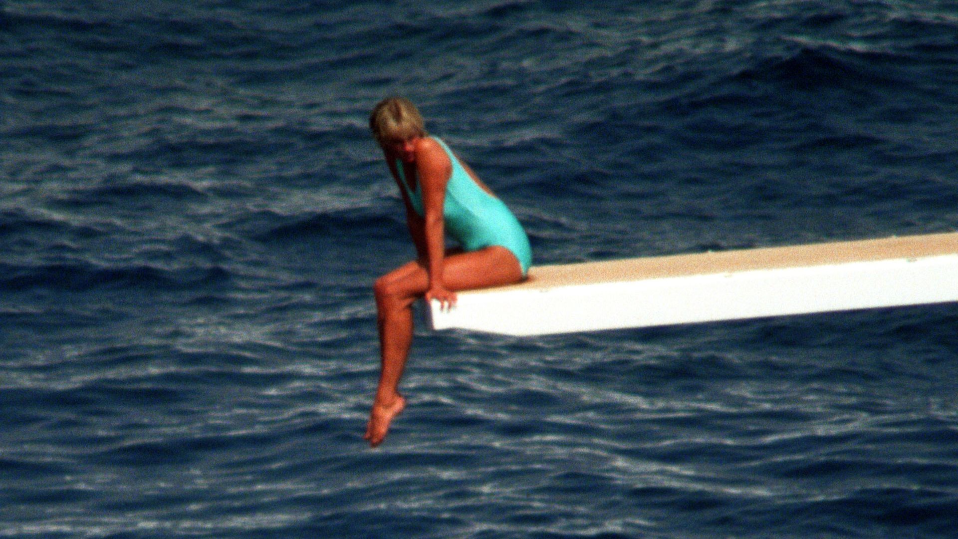 Princess Diana in a blue swimsuit sat on a diving board with the sea beneath her