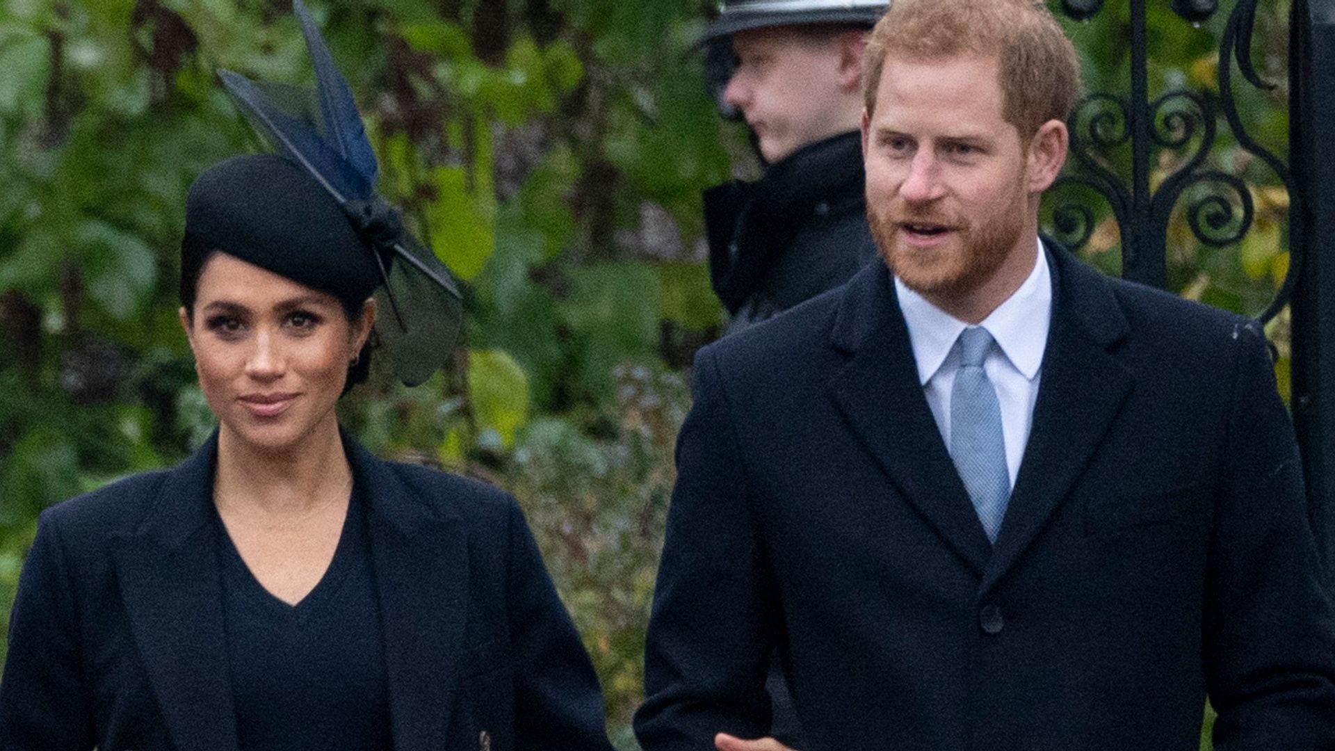 Meghan Markle in a blue hat and coat linking arms with Prince Harry in front of a metal gate