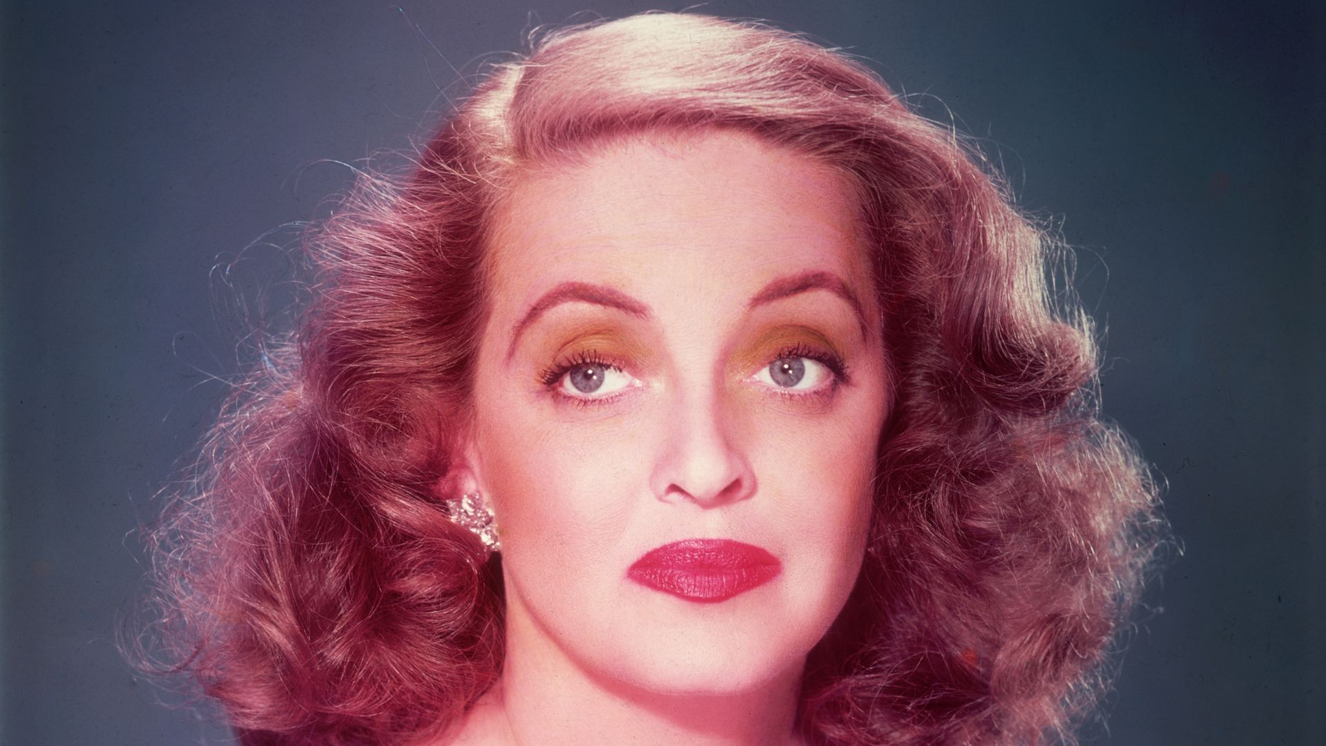 Promotional studio portrait of American actor Bette Davis (1908 - 1989) wearing a violet off-shoulder gown with a jeweled brooch against a blue background, for director Joseph Mankiewitz's film, 'All About Eve'