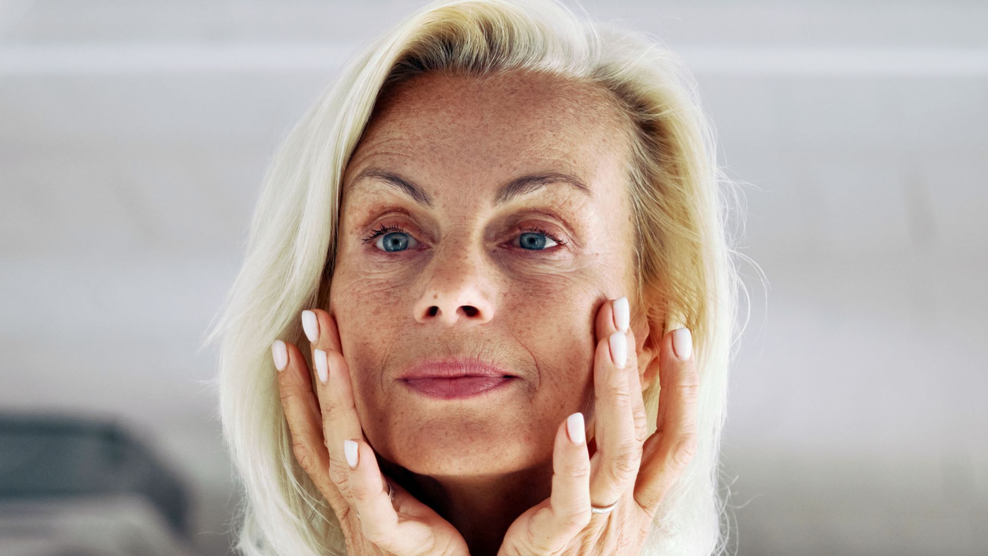 Close up of older 65s woman stand in front of mirror, gently touch her face with hands, examining skin, assessing results of skincare routine.