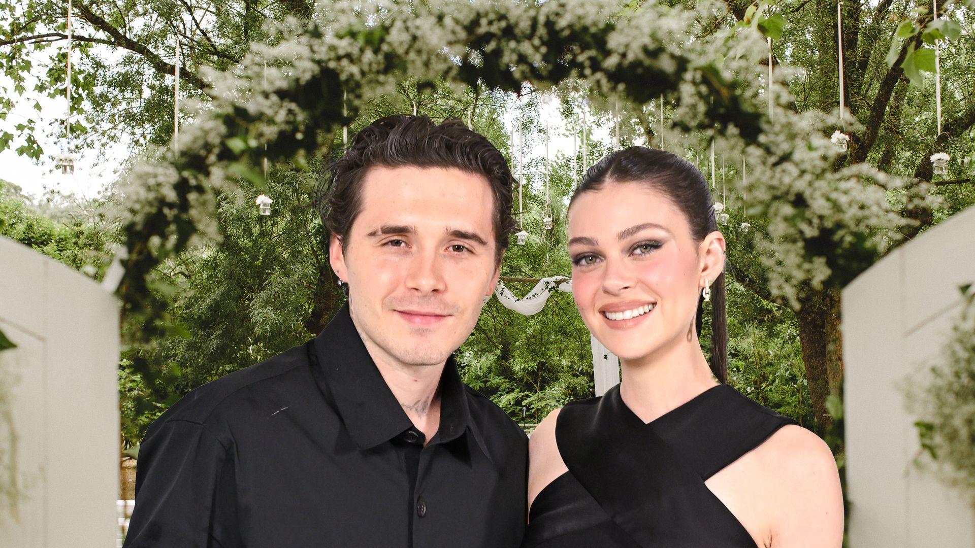 Brooklyn and Nicola Peltz both smiling, dressed in black in front of a rustic boho wedding ceremony set up