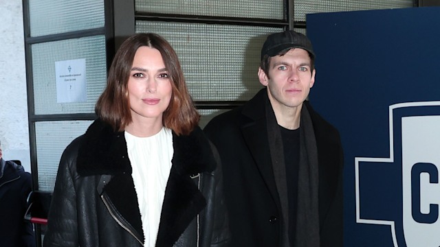 Keira Knightley walks hand-in-hand with James Righton past a "1907 COMO" football logo. She shines in her artistic white Erdem dress, toughened up by a black leather jacket and boots. James contrasts with a black overcoat and cap.