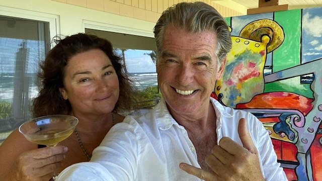 Pierce Brosnan smiling in a white shirt next to his wife holding a drink