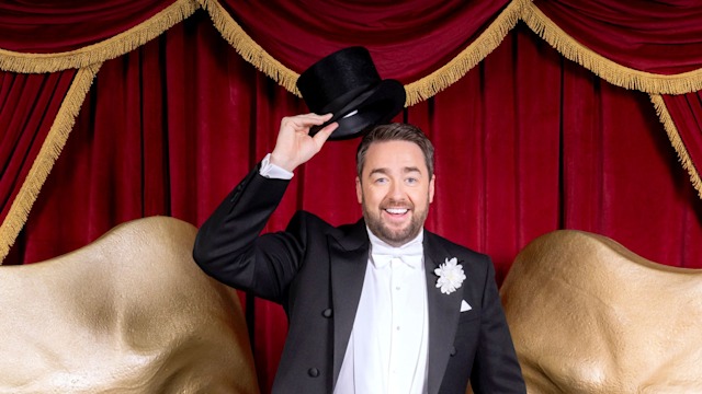 Jason Manford in a top hat and tailsuit with a cane in front of two curtains