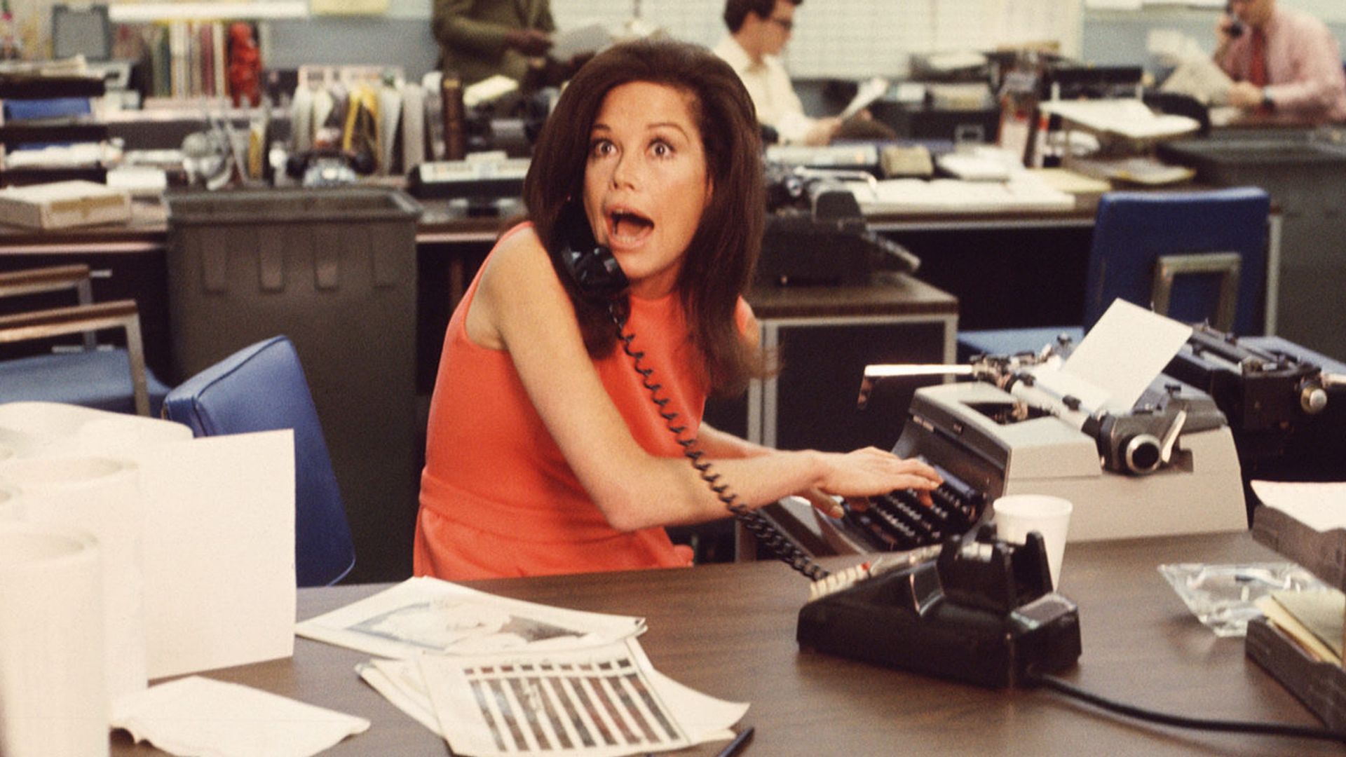 American actress Mary Tyler Moore mouths surprise on the telephone while simultaneously typing as others work in the background in a scene from 'The Mary Tyler Moore Show' (also known as 'Mary Tyler Moore'), Los Angeles, California, early 1970s. Moore wears a sleevless orange dress as she sits behind her desk. (Photo by CBS Photo Archive/Getty Images)