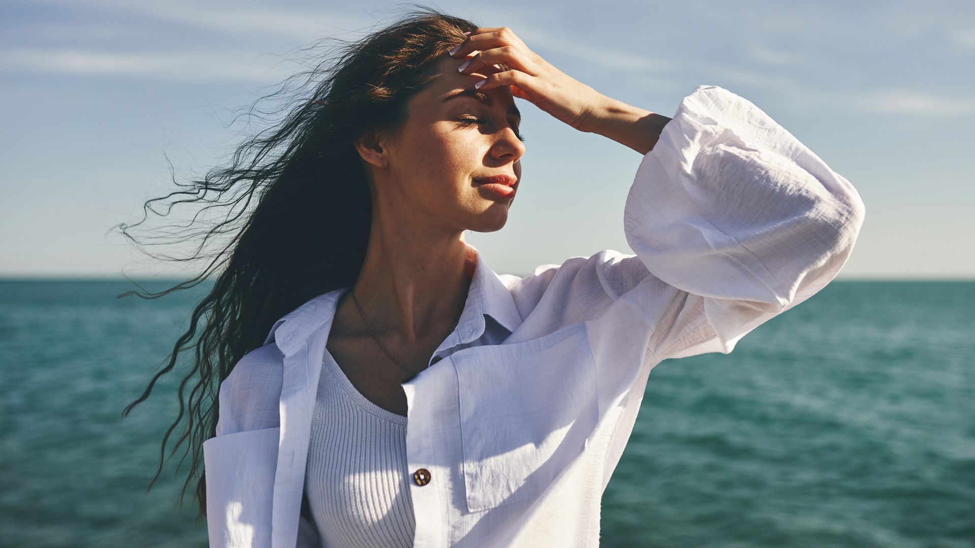 A woman standing by the sea with her hand on her forehead, looking thoughtful. Her hair is blowing in the wind. The sky is clear and the sea is in the background.