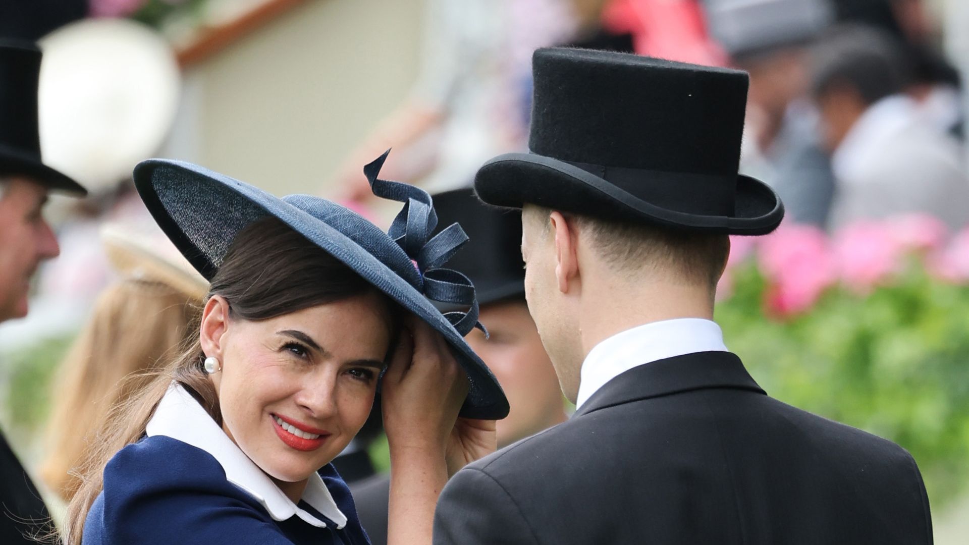 Sophie Winkleman adjusting her hat as Lord Frederik Windsor holds her bag