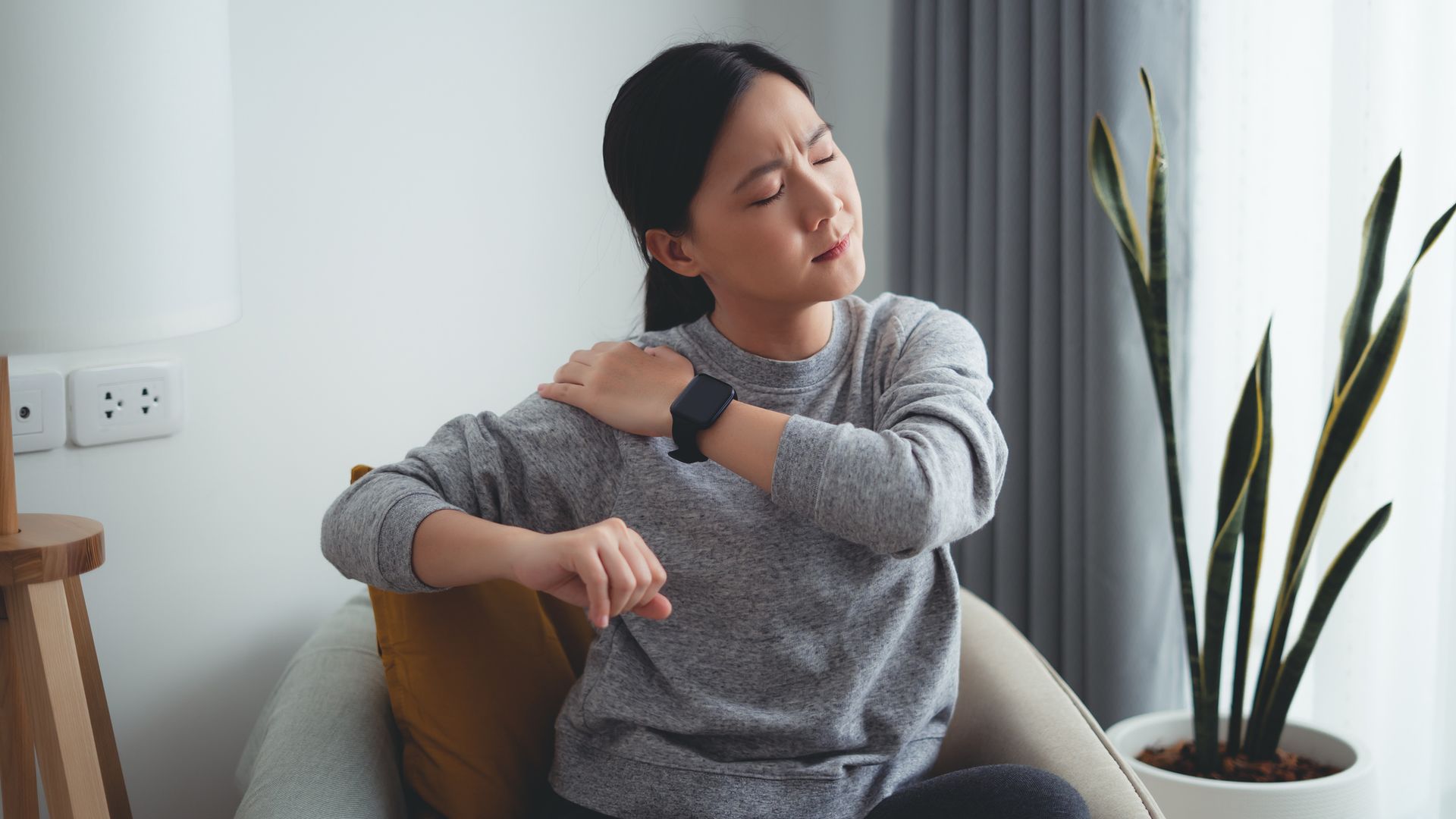 Asian woman suffering from shoulder pain massaging on shoulder by hands sitting on armchair in living room at home.