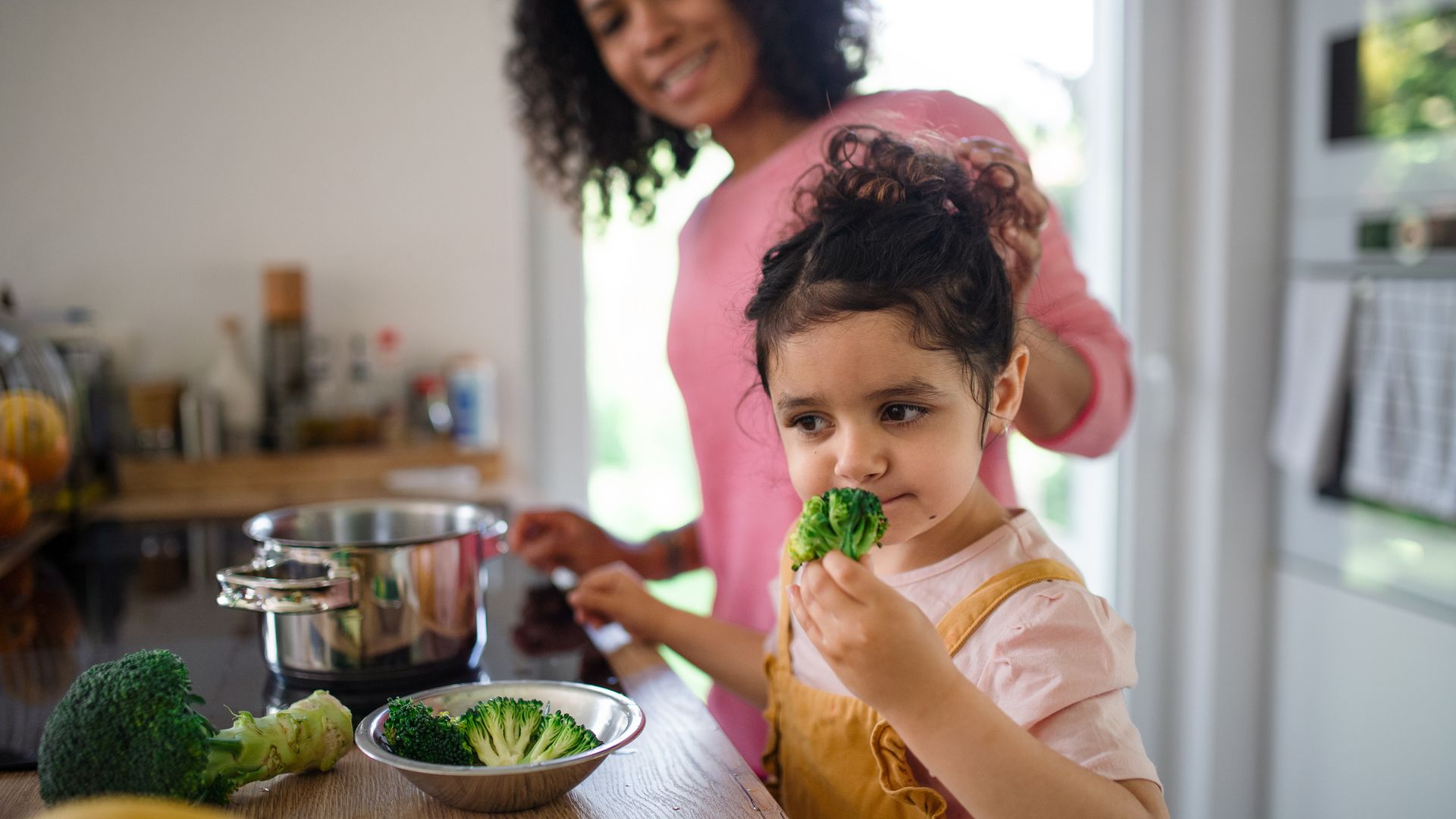 Happy girl preparing broccoli for lunch with her mother in kitchen.