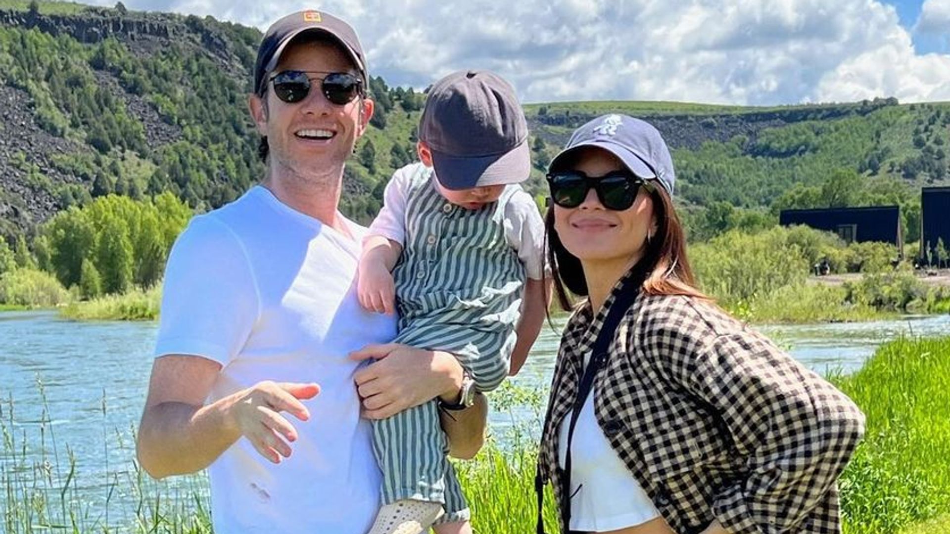 Olivia Munn and John Mulaney smiling in a field near a river with their son Malcolm in John's arms