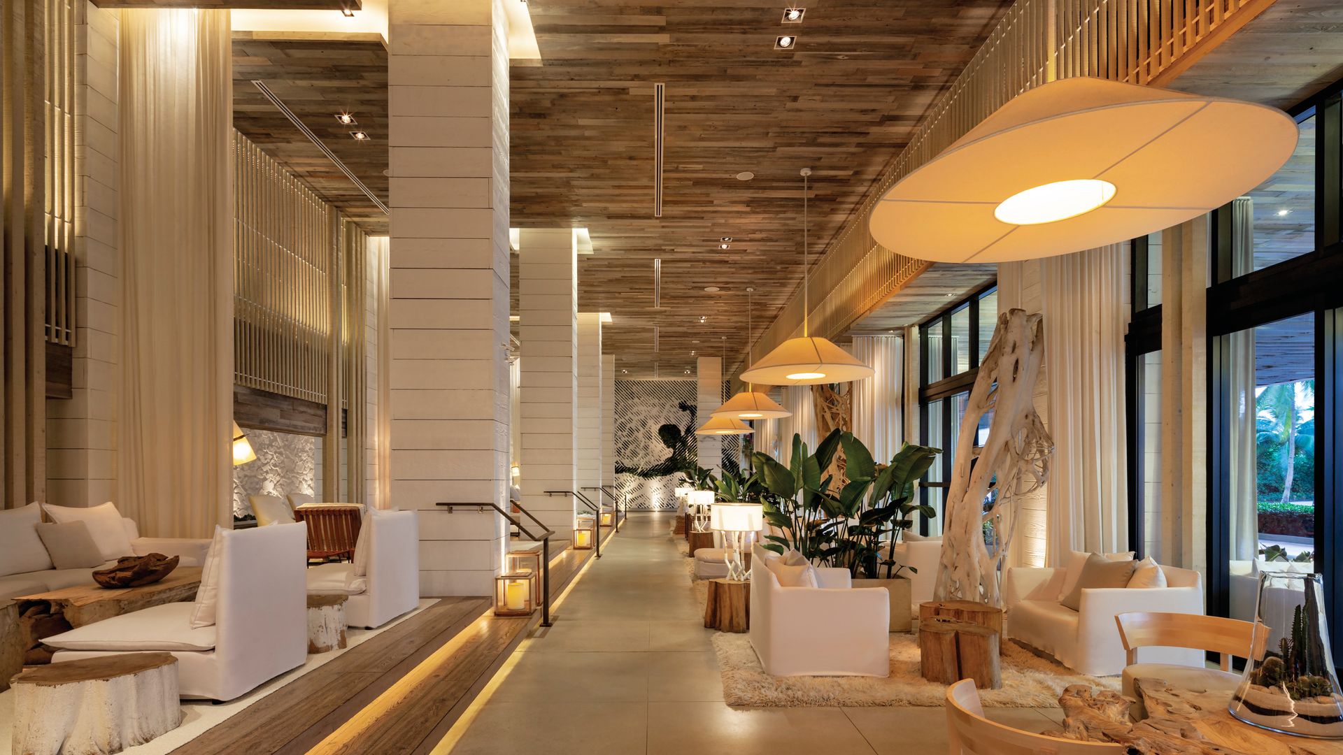 A sophisticated, nature-inspired hotel lobby at 1 Hotel South Beach featuring neutral-toned furniture, organic textures, and reclaimed wood accents. Soft white seating is arranged around driftwood coffee tables, while oversized pendant lights cast a warm glow.