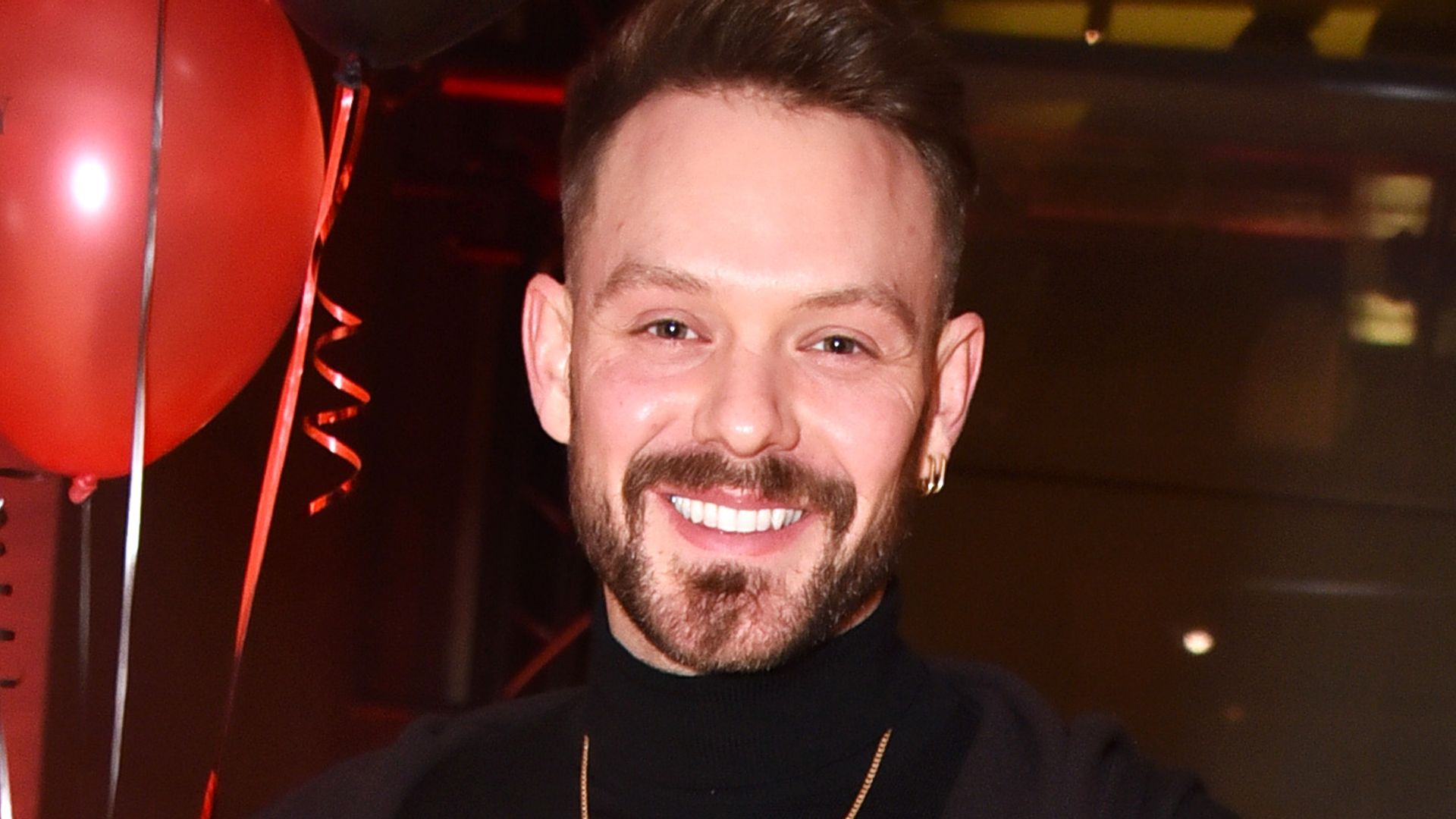John Whaite in a black outfit in front of red balloons