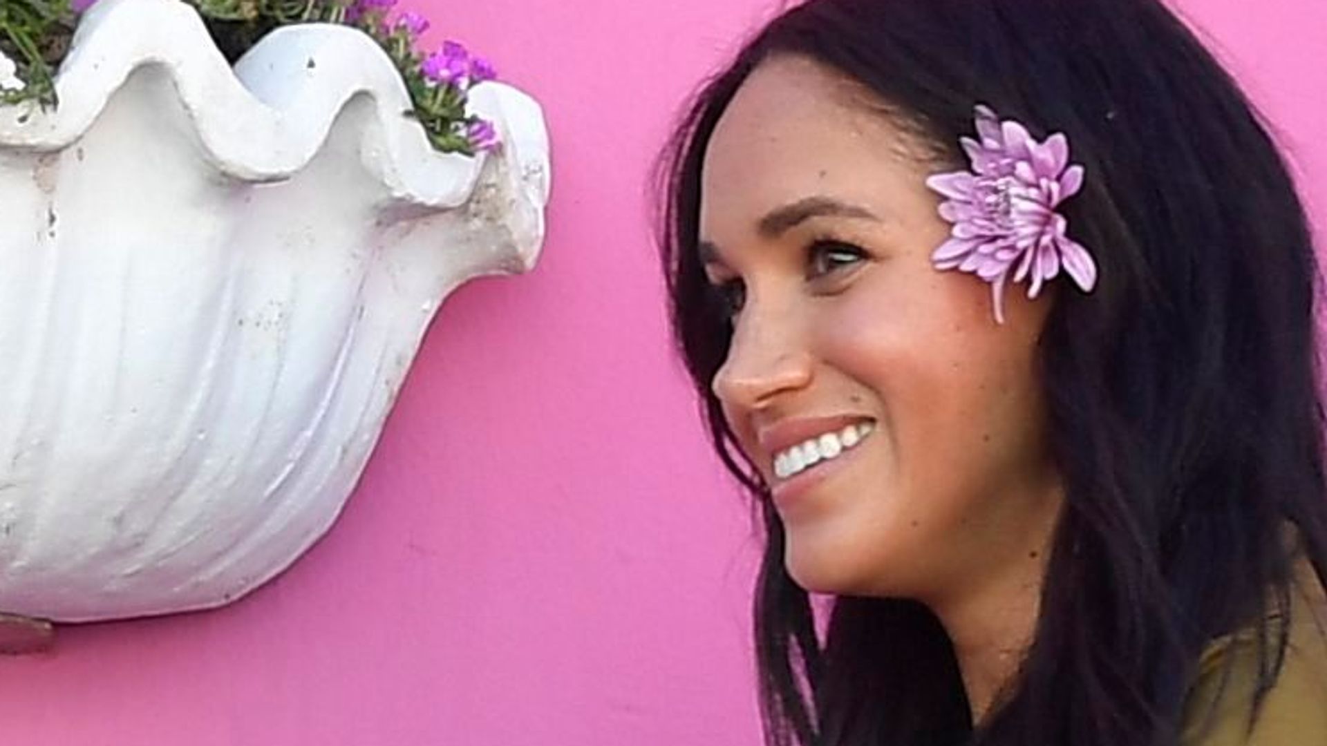 The Duchess of Sussex in front of a pink wall with a flower in her hair