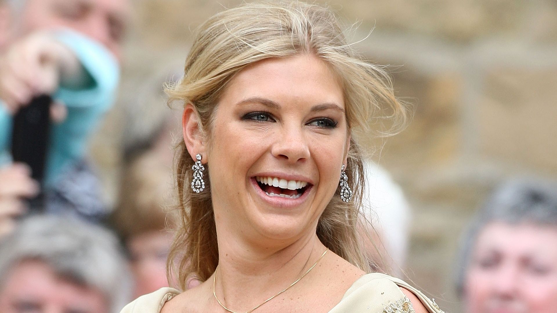 Chelsy Davy smiling in a gold dress and drop earrings at Melissa Percy's wedding