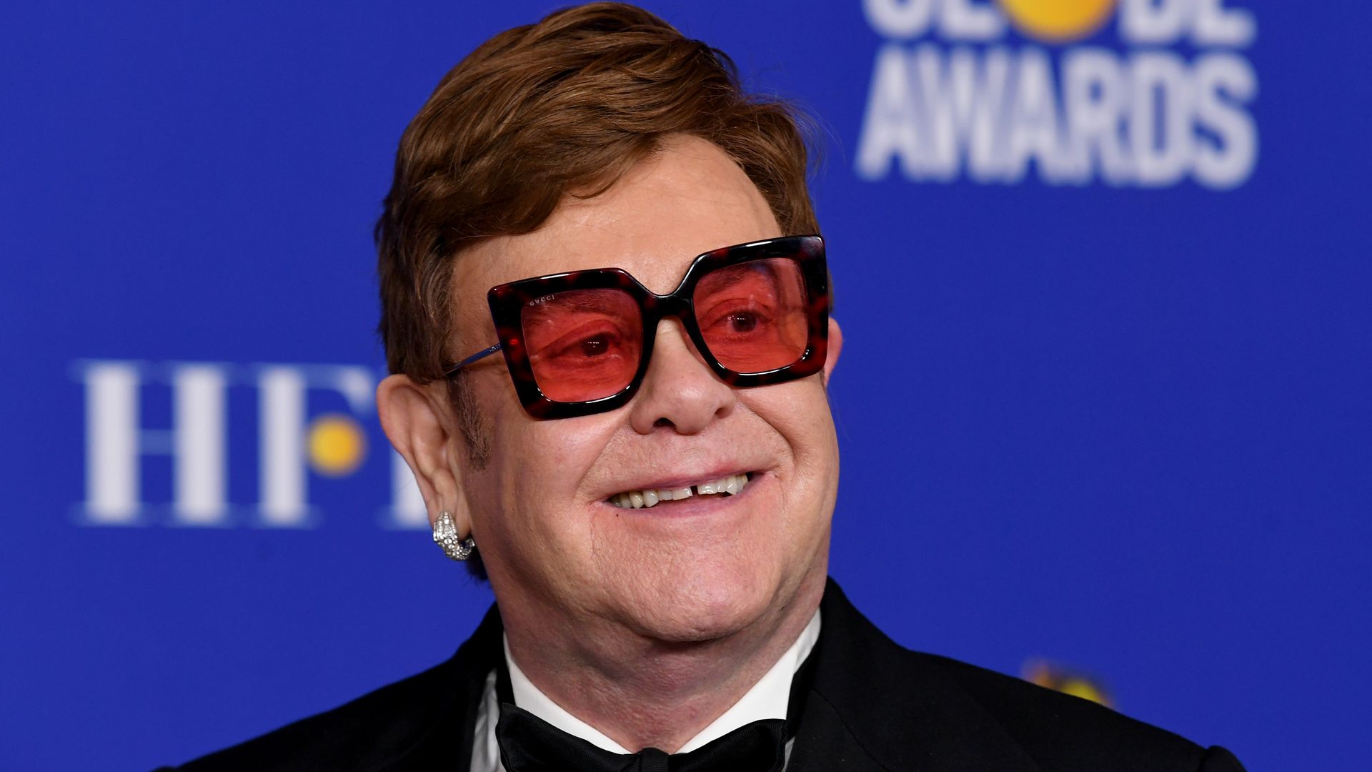 Sir Elton John wearing red-tinted glasses and a tuxedo while walking red carpet at golden globe awards