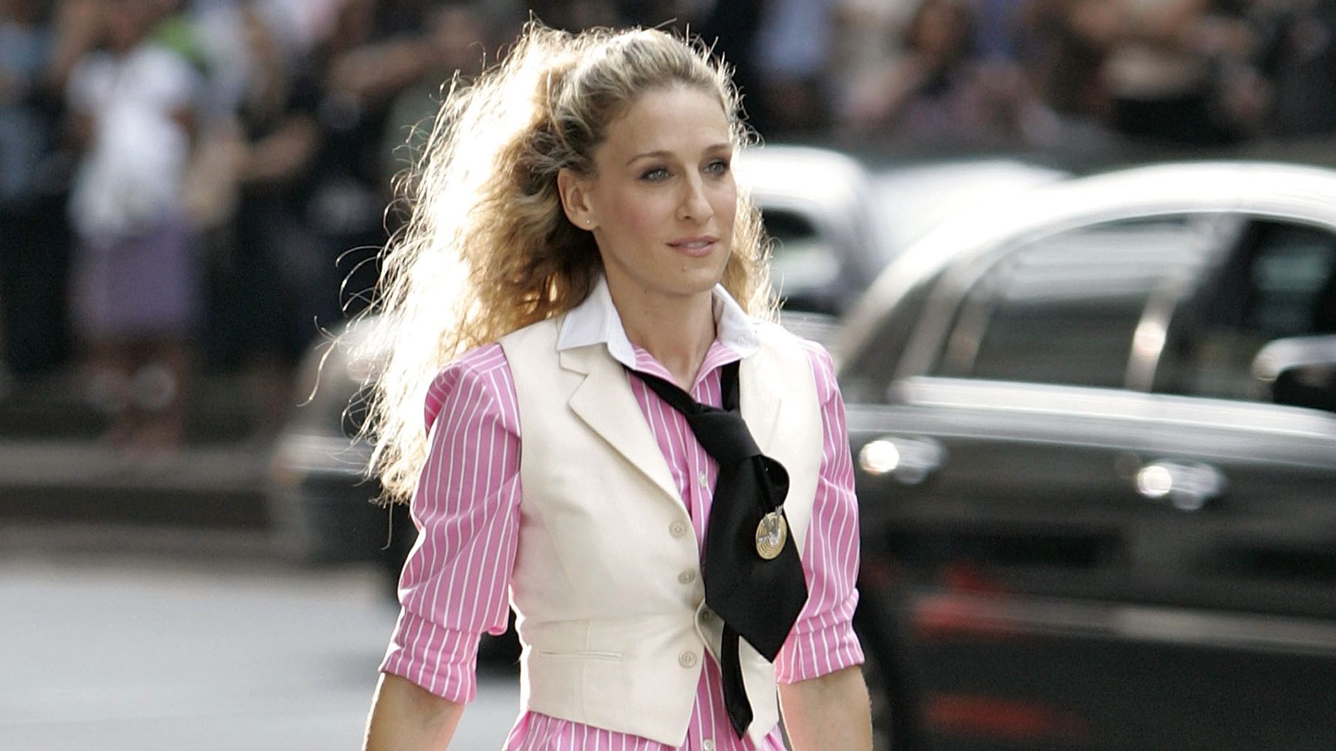 Carrie Bradshaw wearing a cream waistcoat, black tie, pink striped shirt, tailored trousers and holding a round hat bag  