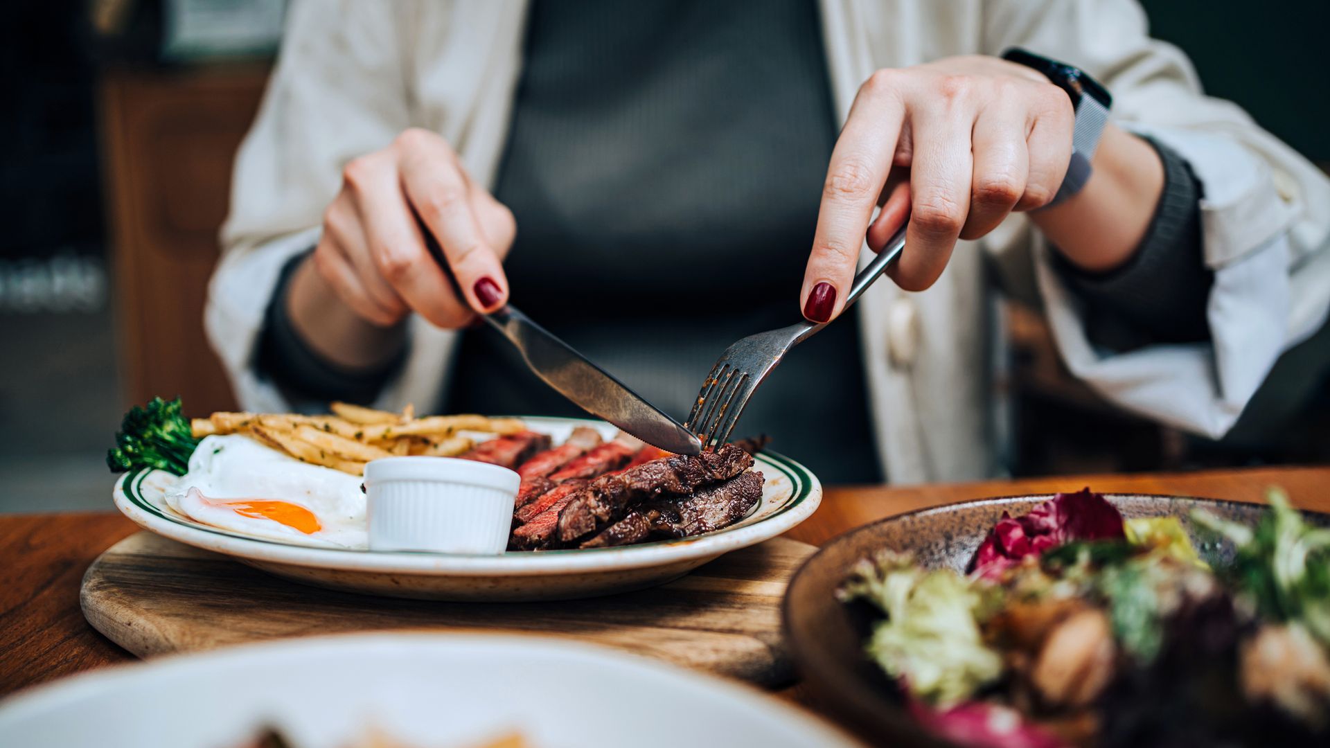 Close-up, mid-section of young Asian woman eating a juicy, grilled fillet steak on her plate along with assorted dishes on dining table, enjoying her meal in an outdoor restaurant. Outdoor dining. People, lifestyle and food concept