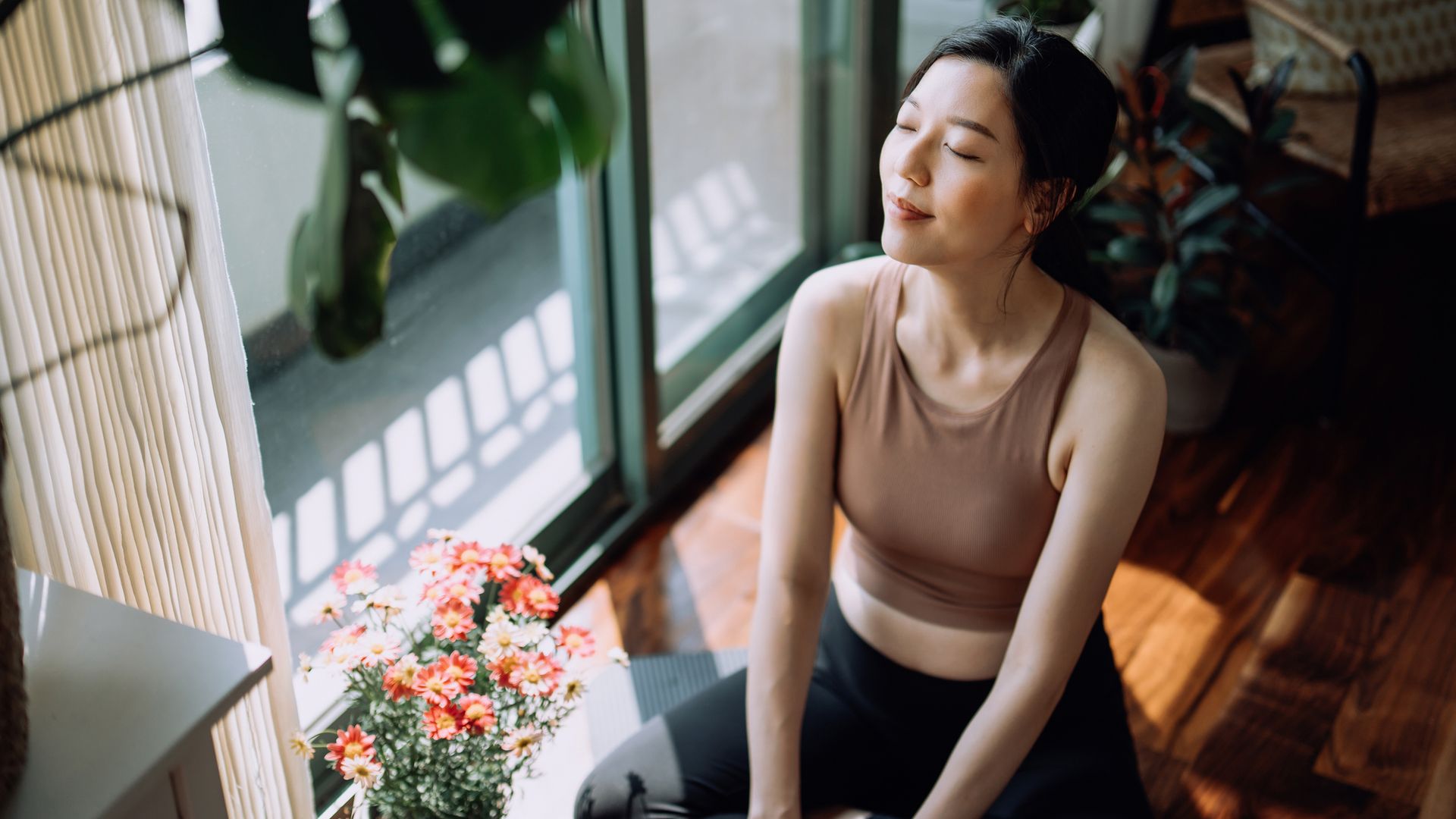 Active young Asian sports woman taking a break after working out at home, sitting on exercise mat taking a deep breath with her eyes closed. Sports and exercise routine. Health, fitness and wellness concept
