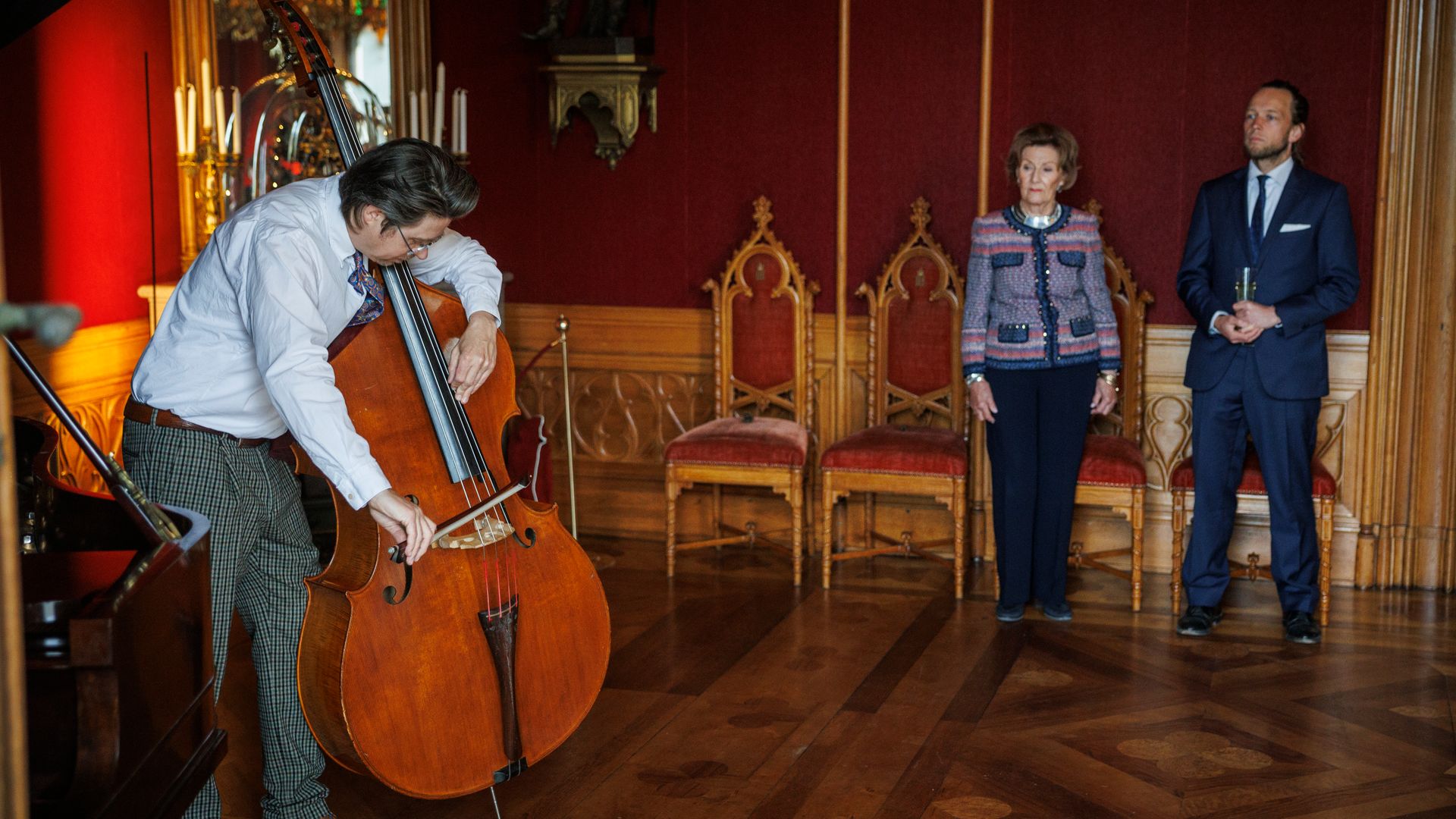 Queen Sonja and a man inside an ornate room watching a man play the cello