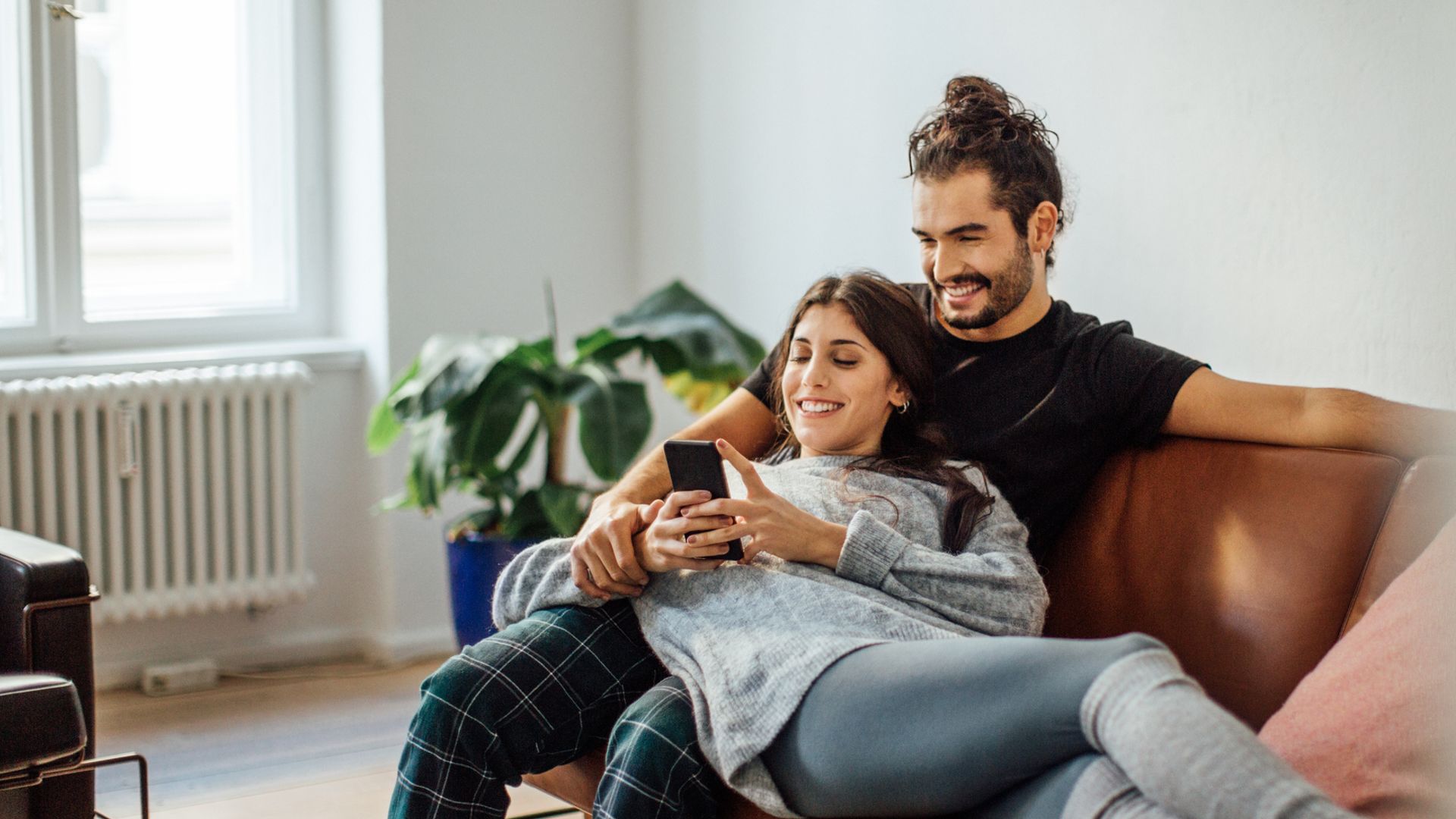 Smiling young woman using mobile phone while reclining on boyfriend sitting on sofa at home