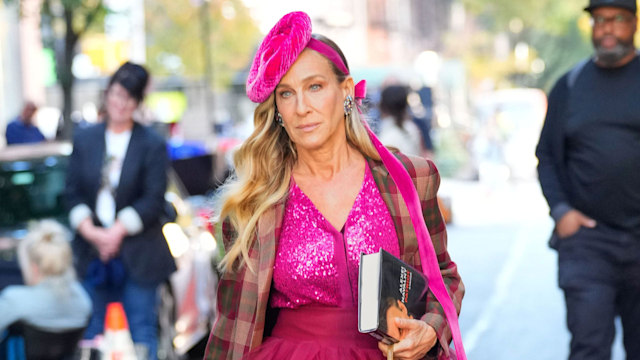 Carrie Bradshaw wears a vibrant pink sequined top with a voluminous magenta tulle skirt. She layers a plaid, oversized coat for contrast. Accessories include a bright pink fascinator, oversized earrings, a tan woven purse, and hot pink strappy heels. She holds a book, exuding confidence and signature bold style.