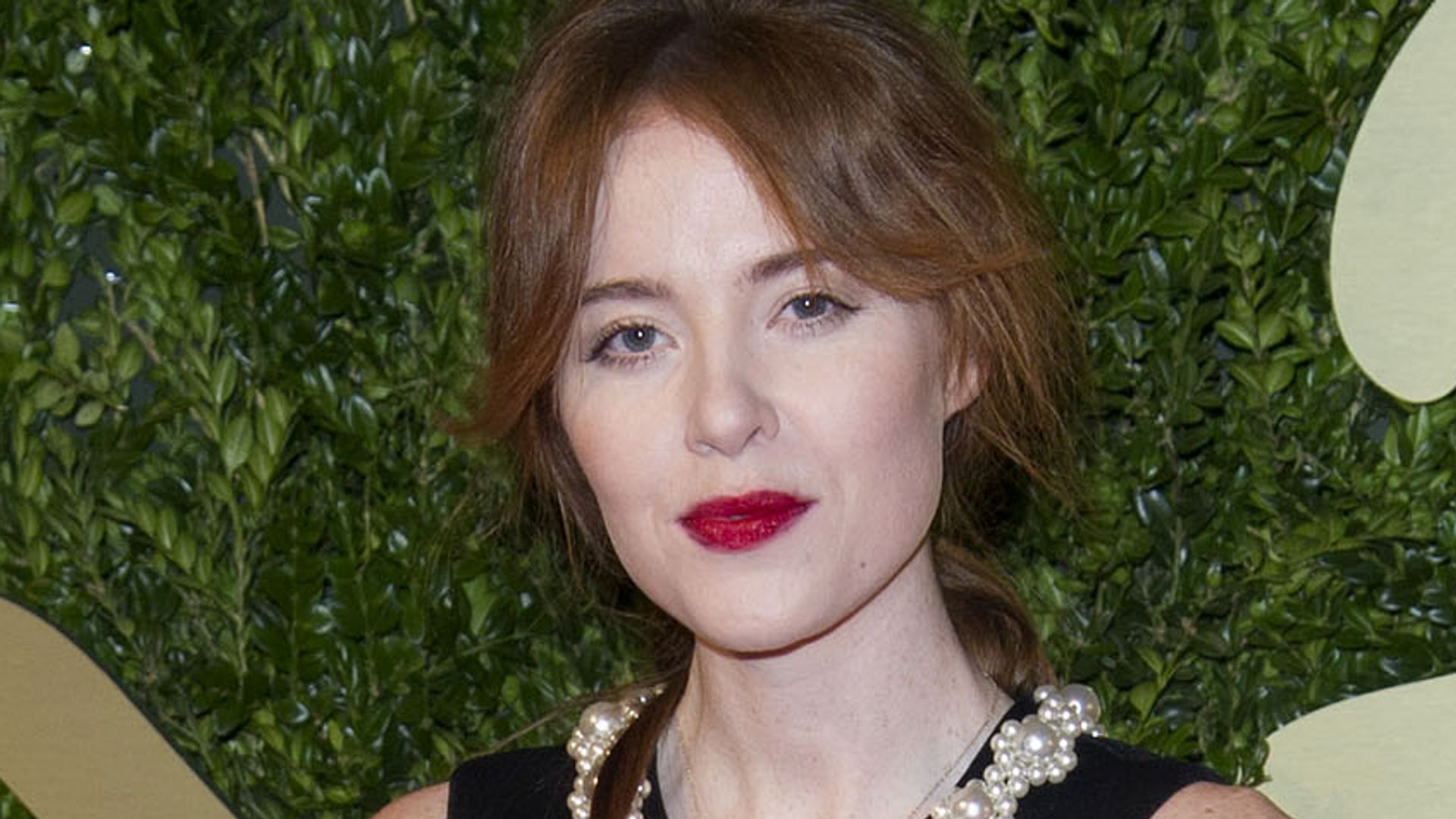 Angela Scanlon in a black dress and red lipstick against a leafy backdrop