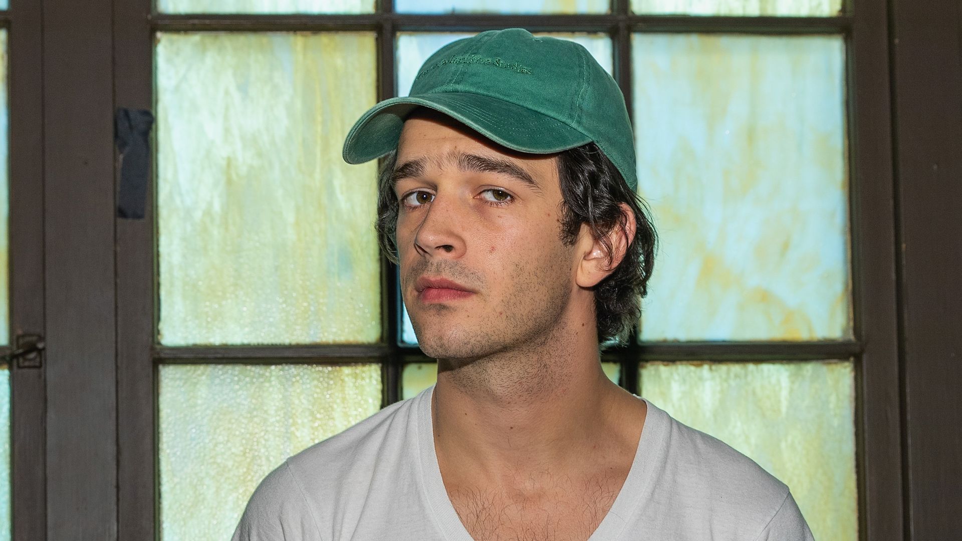 Matty Healy of the 1975 poses for a photo in a white tee and green cap